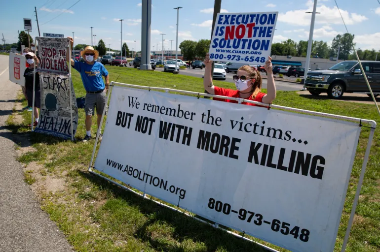 protesters hold signs against the death penalty outside of a federal execution facility
