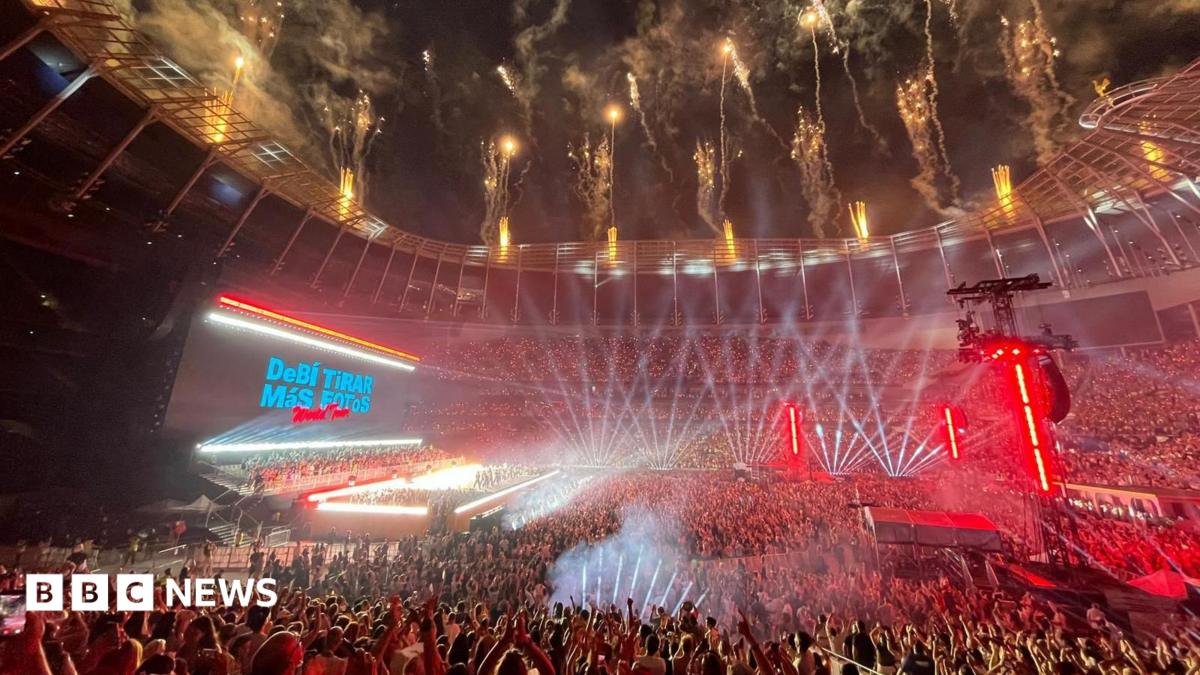 A huge crowd fills the Tottenham Hotspur Stadium, illuminated by stage lights. Fireworks go off from the roof. A stage is lit brightly. Behind, the big screen says Debí Tirar Más Fotos World Tour.