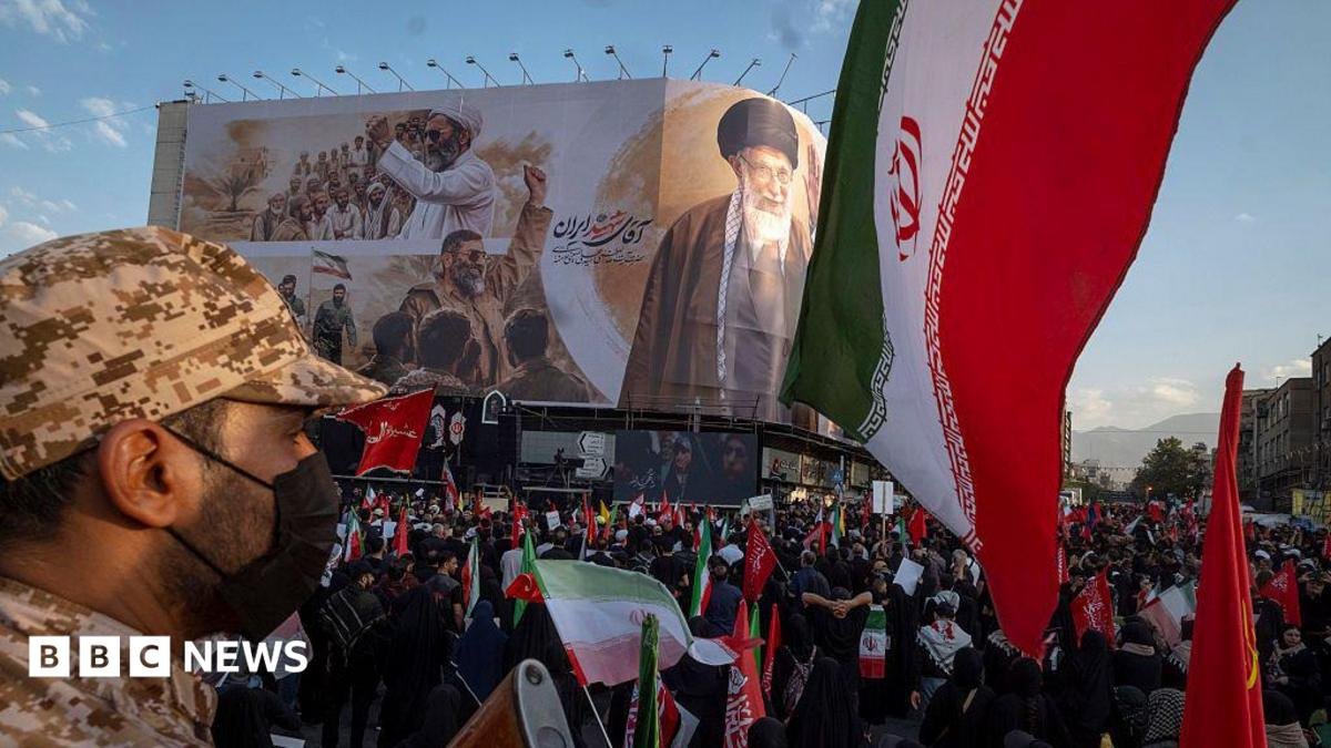 an Iranian security officer amid crowds in front of a billboard showing the late supreme leader at his funeral
