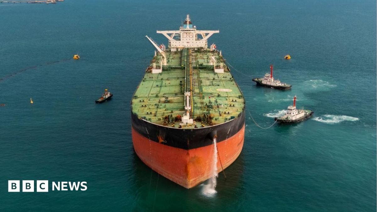 Stock image of an oil tanker pictured front on, with several tugs surrounding the ship