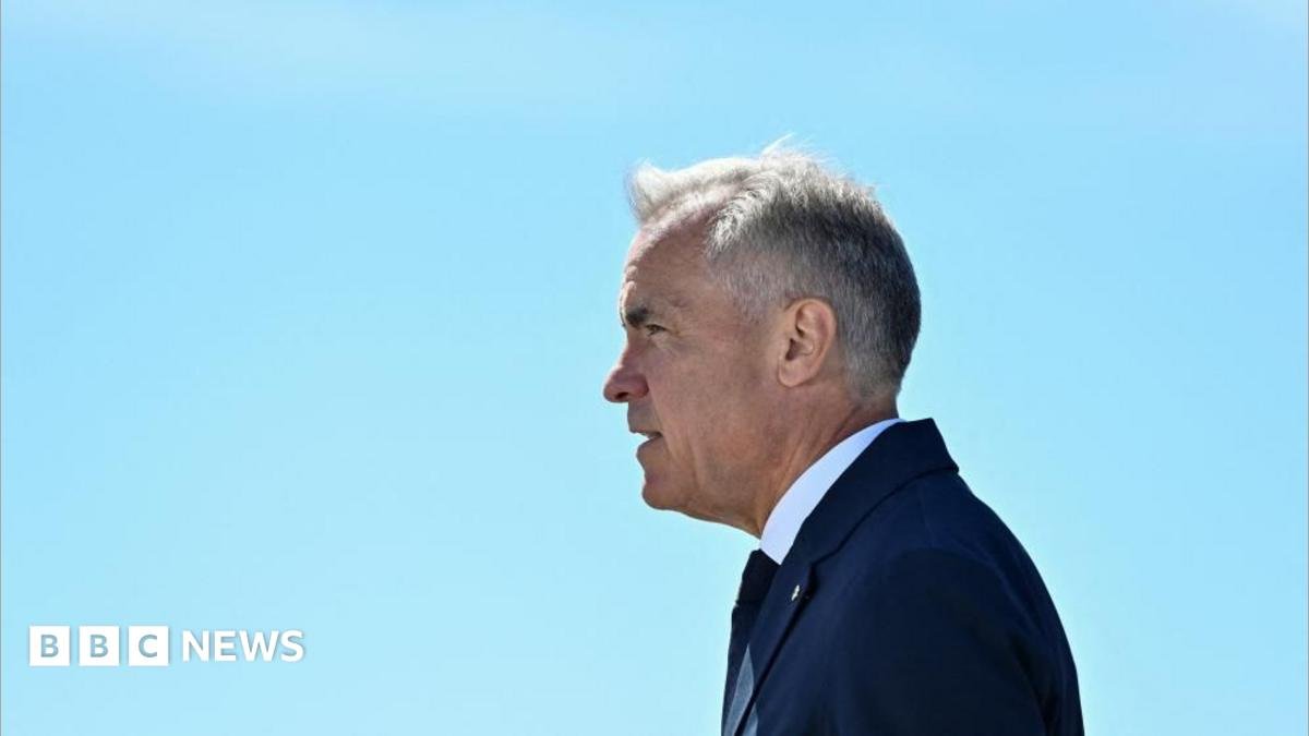 Mark Carney stands in profile, facing to the left, against a clear light blue sky. He wears a dark blue suit and tie.