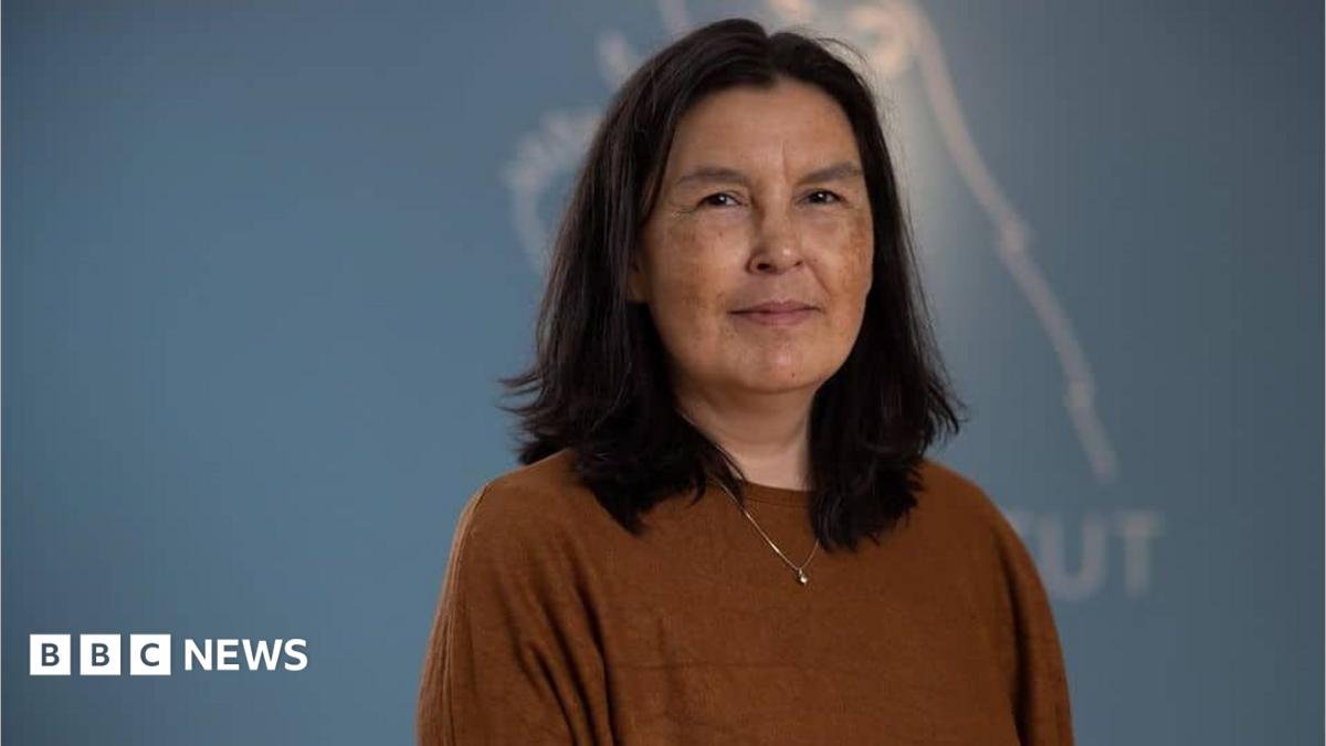 A woman wearing a brown top stands against a blue background