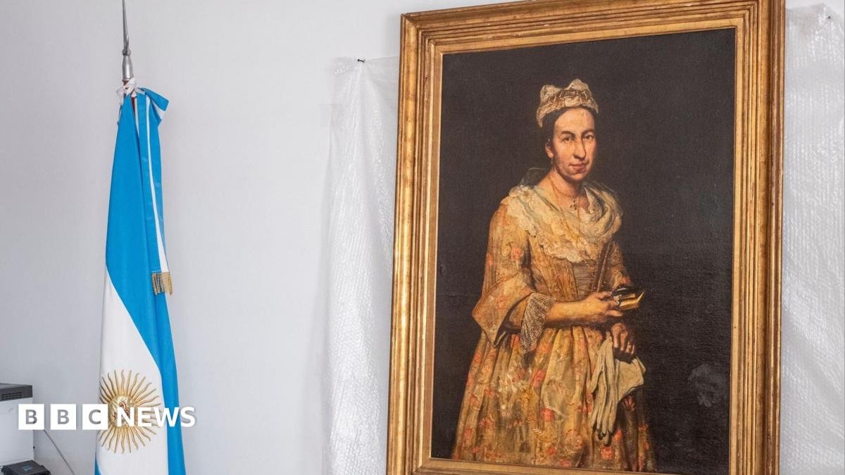 A portrait hangs on the wall with a gold frame on a white backdrop, next to it is an Argentinian flag rolled up