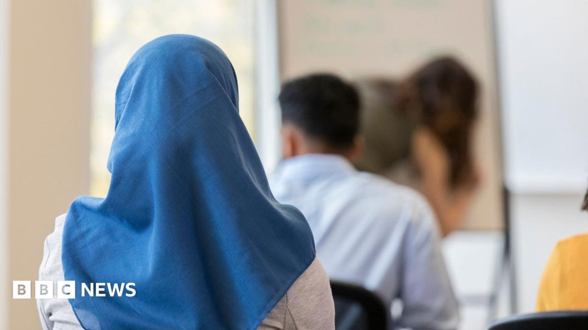 A stock image of a girl wearing a blue headscarf. She is sitting down in a classroom, facing with her back towards the camera.