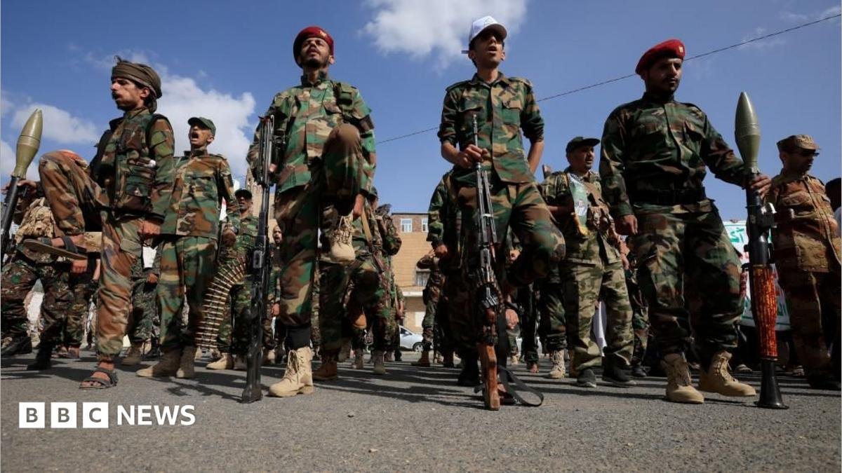 A dozen Houthi men wearing combat clothes, berets and holding large guns. The sky is blue and the men are looking straight ahead.