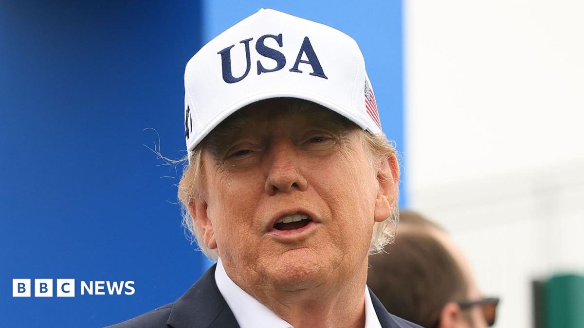 U.S. President Donald Trump attends the Amgen Irish Open at Trump International Golf Links in Doonbeg, Ireland. He is wearing a white USA baseball cap, navy blazer with a white shirt. He has his arms outstretched.