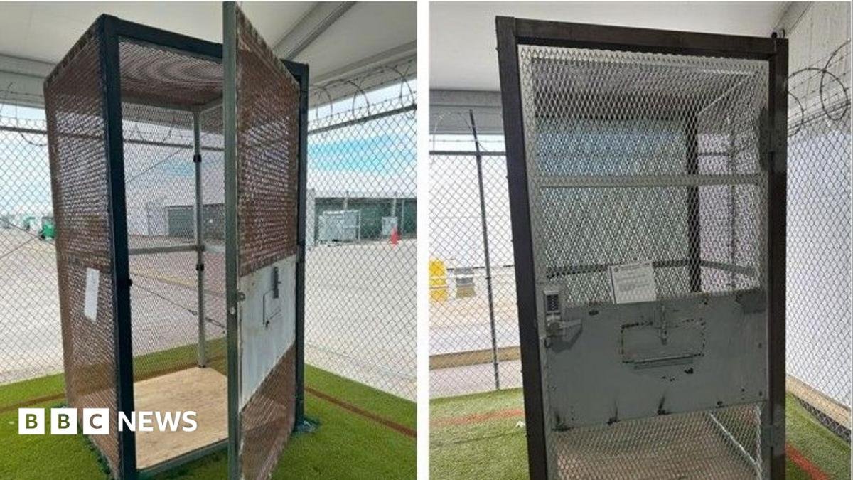 A metal cage sits within a larger cage that has razor wire across the top. The small cage has a door with a combination lock