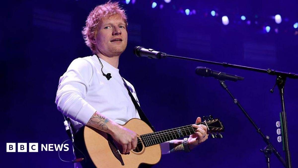 Ed Sheeran, in a long sleeved white T-shirt, holds a guitar and sings into a microphone. He is looking slightly off to one side.