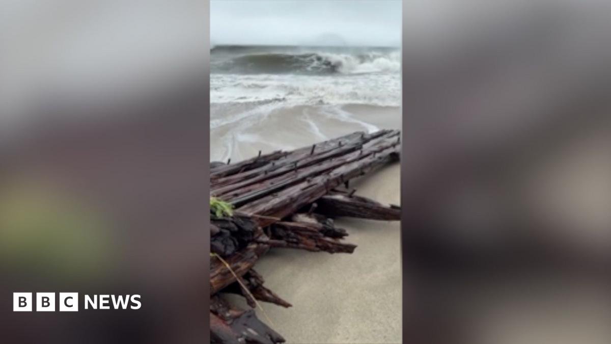 A wooden shipwreck washed up on a beach with big waves in the background.
