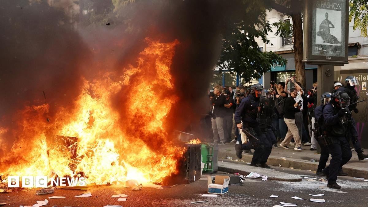 A large fire burns on a road in Paris beside which five officers in riot gear walk past a crowd observing the flames. There is debris strewn across the street.