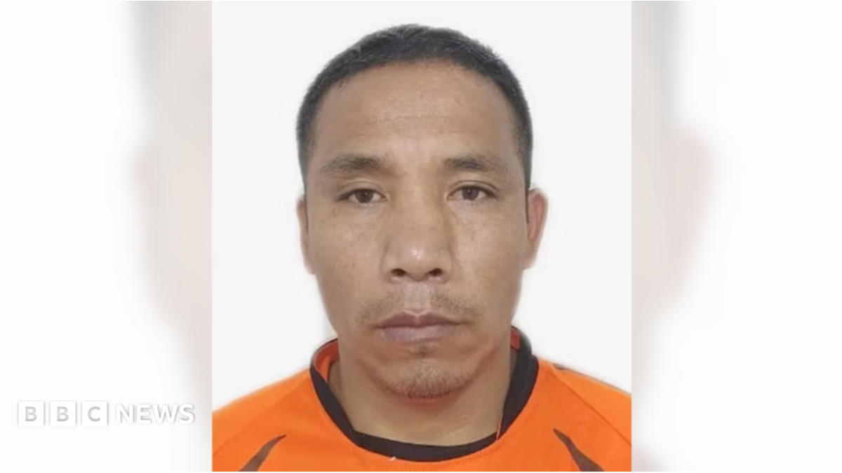 A man pictured against a white background. He is middle aged with cropped black hair and wears an orange and black top.
