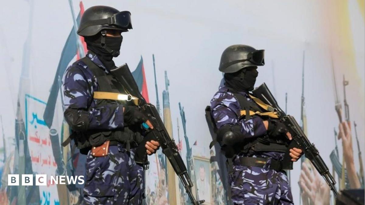 Two soldiers with guns in blue camouflage standing guard in front of a big mural that has may pictures of guns on it.